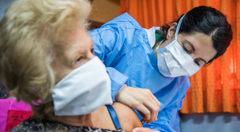 Una trabajadora sanitaria vacuna a una persona mayor, ambas con barbijo.