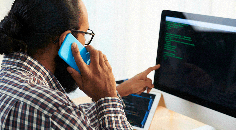 Hombre sentado sostiene un teléfono celular y apunta a su monitor. 