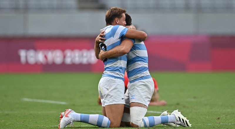 Dos jugadores de rugby de Argentina, arrodillados en el césped, se abrazan.