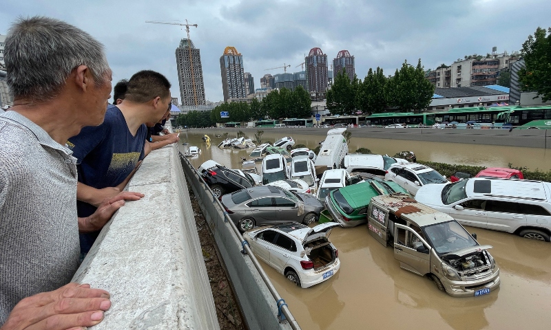 Personas miran desde un puente autos que flotan en medio de una inundación.