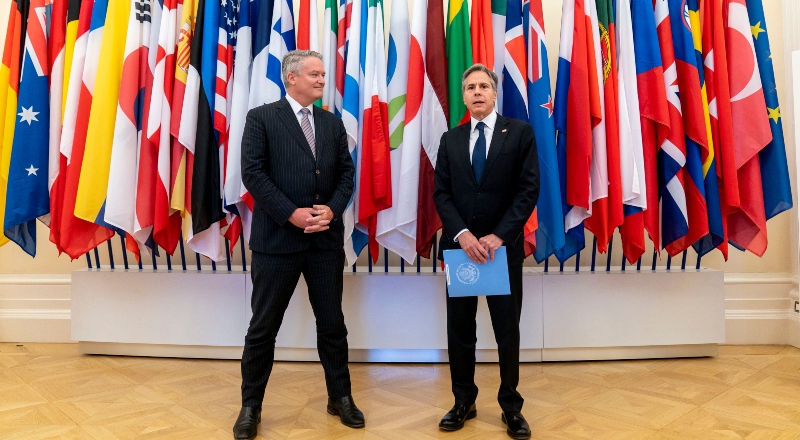 Dos hombres de traje dialogan en una reunión de líderes internacional, con banderas de fondo.